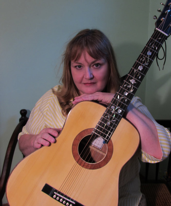 Diane Kauffmann seated with an acoustic guitar she built, resting her arms across the body. The fretboard is inlaid with a mother-of-pearl floral vine.